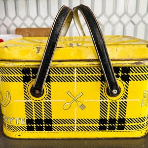 May include: Vintage yellow metal picnic basket with black plaid pattern. Features a black handle and a design of a spoon and fork. The basket has a rectangular shape and is likely used for carrying food and drinks.