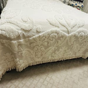 May include: A white bedspread with a floral pattern and a fringed edge. The bedspread is made of a soft, textured fabric and covers the bed. The overall theme is bedding and home decor.