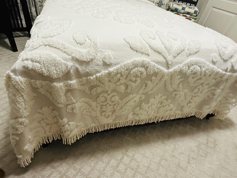 May include: A white bedspread with a floral pattern and a fringed edge. The bedspread is made of a soft, textured fabric and covers the bed. The overall theme is bedding and home decor.