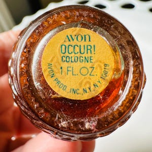 K&ouml;nnte beinhalten: Nahaufnahme einer Avon Occur! Cologne-Flasche. Die braune Flasche hat ein strukturiertes Design. Ein gelbes Etikett auf der Unterseite lautet "AVON OCCUR! COLOGNE 1 FL. OZ." in gr&uuml;ner Schrift. Die Flasche wird von einer Person gehalten.