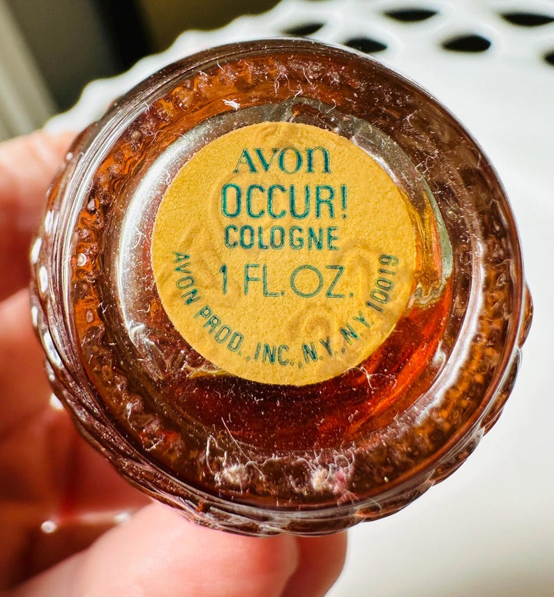 K&ouml;nnte beinhalten: Nahaufnahme einer Avon Occur! Cologne-Flasche. Die braune Flasche hat ein strukturiertes Design. Ein gelbes Etikett auf der Unterseite lautet "AVON OCCUR! COLOGNE 1 FL. OZ." in gr&uuml;ner Schrift. Die Flasche wird von einer Person gehalten.