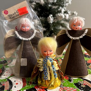 May include: Three vintage angel ornaments with blonde hair and fabric wings. One angel is in a plastic bag with a price tag. The angels are on a Christmas-themed tablecloth.