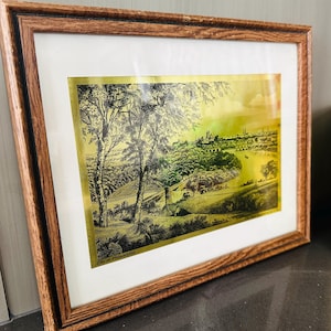 May include: Framed vintage print of a landscape scene with trees, a river, and buildings. The print has a gold-toned background and is framed in a brown wooden frame. The text "City of Philadelphia" is visible at the bottom.
