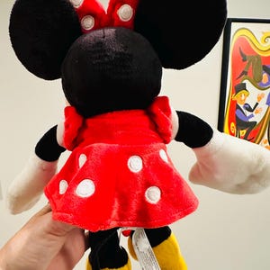 Puede incluir: Un mu&ntilde;eco de peluche de Minnie Mouse, visto desde atr&aacute;s, con un vestido rojo con lunares blancos y un lazo a juego. El mu&ntilde;eco tiene orejas negras, guantes blancos y zapatos amarillos. Una etiqueta est&aacute; adjunta.