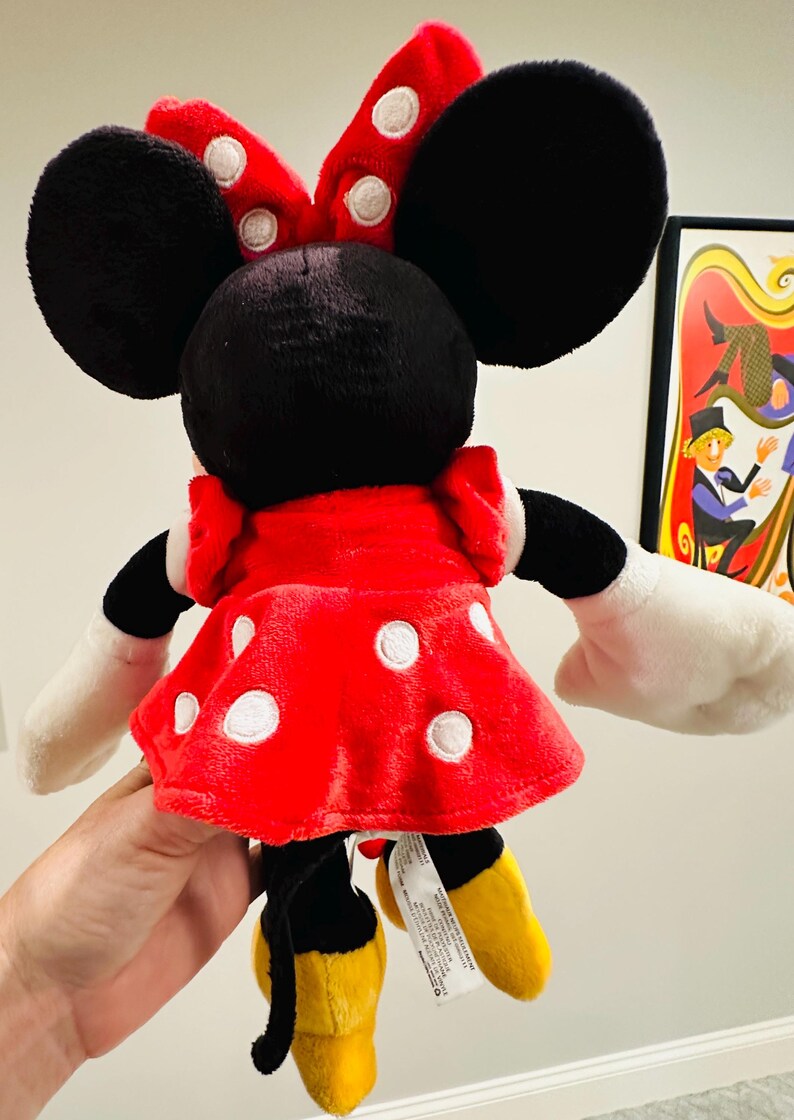 Puede incluir: Un mu&ntilde;eco de peluche de Minnie Mouse, visto desde atr&aacute;s, con un vestido rojo con lunares blancos y un lazo a juego. El mu&ntilde;eco tiene orejas negras, guantes blancos y zapatos amarillos. Una etiqueta est&aacute; adjunta.