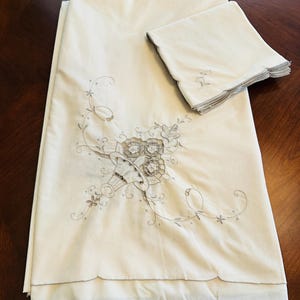 May include: Off-white linen tablecloth and matching napkins. The tablecloth features an embroidered floral design in shades of beige and gray. The napkins have a scalloped edge and a small embroidered detail. The set is folded and displayed on a dark wood surface.