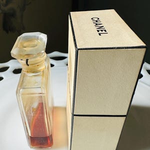 K&ouml;nnte beinhalten: Eine Vintage-Parf&uuml;mflasche mit einem klaren, facettierten Verschluss und einem rechteckigen Glask&ouml;rper. Die Flasche enth&auml;lt eine rotbraune Fl&uuml;ssigkeit. Daneben befindet sich eine beige Schachtel mit dem schwarzen Aufdruck "CHANEL".