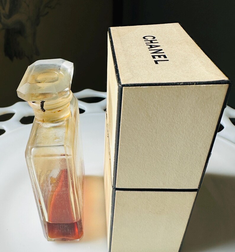 K&ouml;nnte beinhalten: Eine Vintage-Parf&uuml;mflasche mit einem klaren, facettierten Verschluss und einem rechteckigen Glask&ouml;rper. Die Flasche enth&auml;lt eine rotbraune Fl&uuml;ssigkeit. Daneben befindet sich eine beige Schachtel mit dem schwarzen Aufdruck "CHANEL".