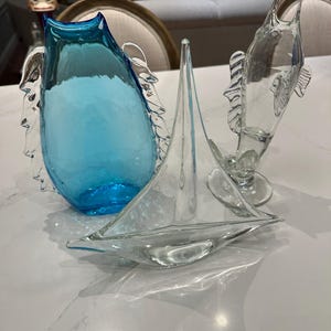 May include: A collection of decorative glass sculptures. Includes a turquoise blue vase with wing-like accents, a clear glass sailboat, and a clear glass fish. The objects are arranged on a white surface.