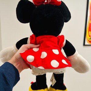 May include: A plush Minnie Mouse doll, viewed from the back, featuring a red dress with white polka dots, a matching bow, and yellow shoes. The doll has black ears and a white lace trim on the undergarment.
