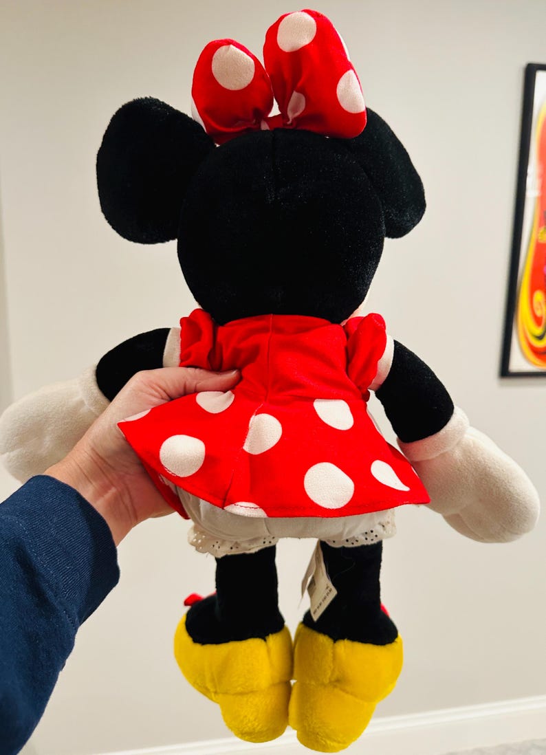 May include: A plush Minnie Mouse doll, viewed from the back, featuring a red dress with white polka dots, a matching bow, and yellow shoes. The doll has black ears and a white lace trim on the undergarment.