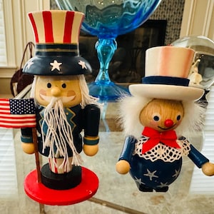 May include: Two wooden patriotic figurines. One is Uncle Sam with a red, white, and blue hat and an American flag. The other is a clown-like figure with a red bow tie and a white hat. Both are on a glass surface.