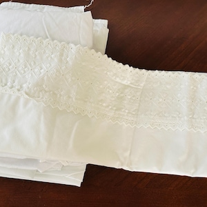 May include: White bedding set featuring a pillowcase with intricate lace detailing and a folded sheet. The pillowcase has a decorative lace border. The sheets are folded neatly on a dark brown surface.