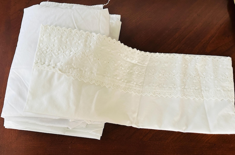 May include: White bedding set featuring a pillowcase with intricate lace detailing and a folded sheet. The pillowcase has a decorative lace border. The sheets are folded neatly on a dark brown surface.