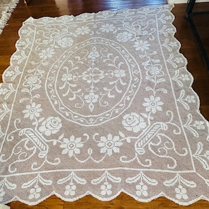 May include: A rectangular white lace tablecloth featuring an intricate crochet pattern with a central oval motif surrounded by floral designs, rose blossoms, and basket elements. The edges are finished with a scalloped border and floral details.