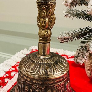 May include: Ornate brass bell with intricate detailing. The bell features a detailed handle with a face and decorative arches. The bell's base has a detailed pattern. The bell is on a red surface.