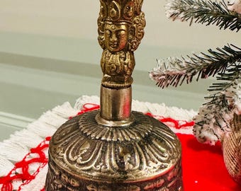 May include: Ornate brass bell with intricate detailing. The bell features a detailed handle with a face and decorative arches. The bell's base has a detailed pattern. The bell is on a red surface.