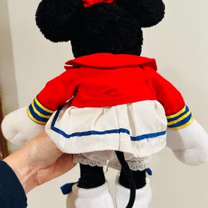 May include: A plush Minnie Mouse doll, viewed from the back, features black ears and a red bow. She wears a red jacket with blue and yellow accents, a white skirt with blue trim, and white gloves and shoes. A black tail is visible.