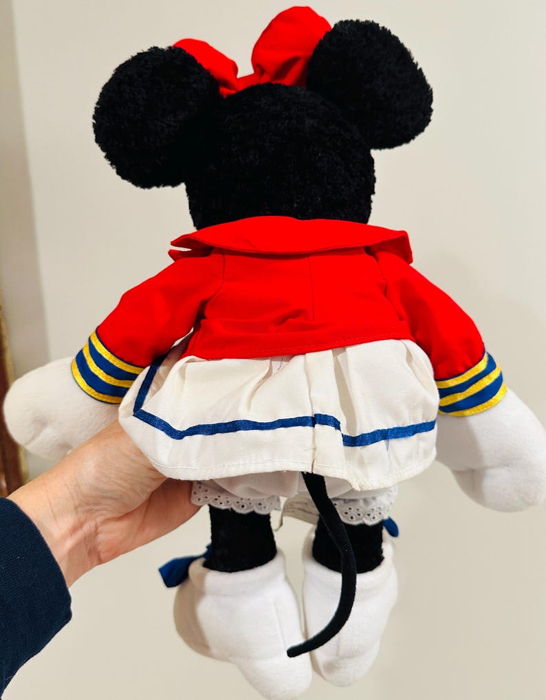 May include: A plush Minnie Mouse doll, viewed from the back, features black ears and a red bow. She wears a red jacket with blue and yellow accents, a white skirt with blue trim, and white gloves and shoes. A black tail is visible.