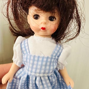 May include: A vintage doll with brown hair and a blue and white checkered dress. The doll has a white blouse and red lips. The doll's hair is styled in a curly fashion. The doll is a collectible item.