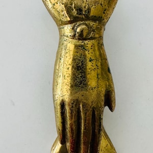 May include: A vintage brass hand-shaped door knocker. The knocker is a detailed sculpture of a hand with a ring at the top for hanging. The brass has an aged, antique appearance.