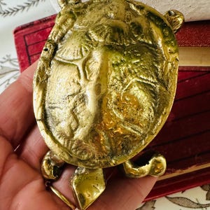 May include: A brass turtle figurine with a detailed shell and four legs. The turtle is a golden color and appears to be vintage. The figurine is held in a hand, with a red book and floral patterned fabric in the background.