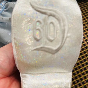 K&ouml;nnte beinhalten: Wei&szlig;er, glitzernder Schuh mit der Zahl 60 und einem stilisierten "D"-Logo auf der Sohle. Der Schuh hat eine weiche, samtige Textur und ist wahrscheinlich ein Gedenkartikel.