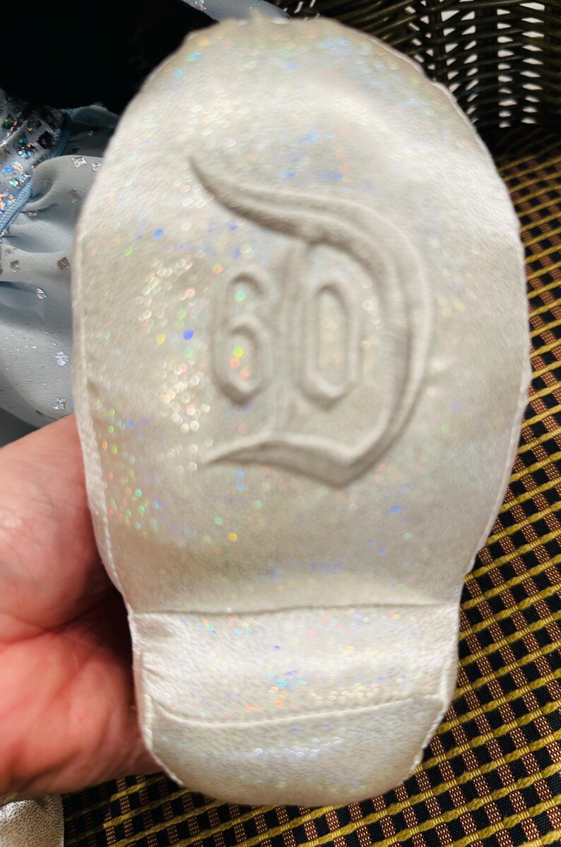 K&ouml;nnte beinhalten: Wei&szlig;er, glitzernder Schuh mit der Zahl 60 und einem stilisierten "D"-Logo auf der Sohle. Der Schuh hat eine weiche, samtige Textur und ist wahrscheinlich ein Gedenkartikel.