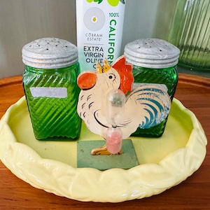 May include: A decorative kitchen set featuring two green glass salt shakers with silver lids, a yellow ceramic tray, and a wooden rooster-shaped timer. The rooster has red, orange, and blue accents. The set is on a wooden surface.