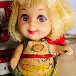 May include: A vintage doll with blonde hair and a smiling expression. The doll wears a small apron with a food graphic and the word "YUM". The doll is a collectible item.