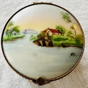 May include: A round, hinged trinket box with a hand-painted landscape scene. The box features a house with a red roof, a bridge, and trees along a body of water. The box has a gold-colored metal rim and a small decorative clasp.