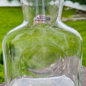 May include: A clear glass decanter with a wide, flat base and a narrow neck featuring a flared rim. A silver-toned sticker on the front displays the text BLENKO HANDCRAFT. The vessel has a distinct circular indentation on the front surface.