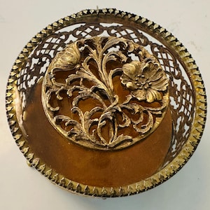 May include: A vintage gold-toned metal sewing pincushion. The round pincushion features a filigree basket design with a floral motif. The center has a brown velvet cushion, and the rim is adorned with a decorative edge.