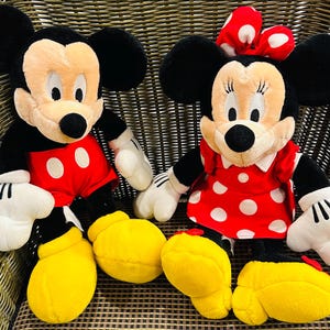 May include: Two plush Disney character dolls: Mickey Mouse and Minnie Mouse. Mickey wears red shorts with white buttons, black ears, and yellow shoes. Minnie wears a red dress with white polka dots and a matching bow.