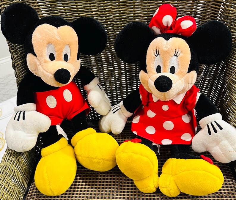 May include: Two plush Disney character dolls: Mickey Mouse and Minnie Mouse. Mickey wears red shorts with white buttons, black ears, and yellow shoes. Minnie wears a red dress with white polka dots and a matching bow.