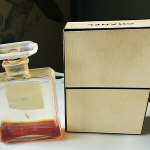 K&ouml;nnte beinhalten: Ein Vintage-Parf&uuml;mflakon mit klarem, geometrischem Design und einem facettierten Verschluss. Die Flasche enth&auml;lt eine rotbraune Fl&uuml;ssigkeit. Eine beige Schachtel mit dem Markennamen "CHANEL" befindet sich neben der Flasche.
