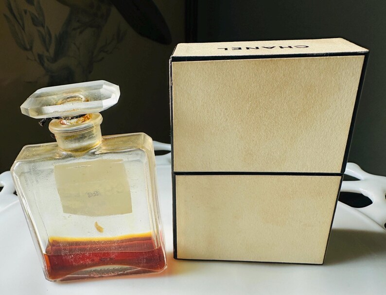 K&ouml;nnte beinhalten: Ein Vintage-Parf&uuml;mflakon mit klarem, geometrischem Design und einem facettierten Verschluss. Die Flasche enth&auml;lt eine rotbraune Fl&uuml;ssigkeit. Eine beige Schachtel mit dem Markennamen "CHANEL" befindet sich neben der Flasche.