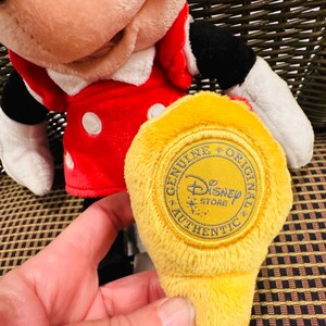Puede incluir: Mu&ntilde;eca de peluche de Minnie Mouse con un vestido rojo con lunares blancos y un cuello blanco. Una etiqueta amarilla con el texto "GENUINE ORIGINAL Disney STORE AUTHENTIC" se sostiene frente a la mu&ntilde;eca.