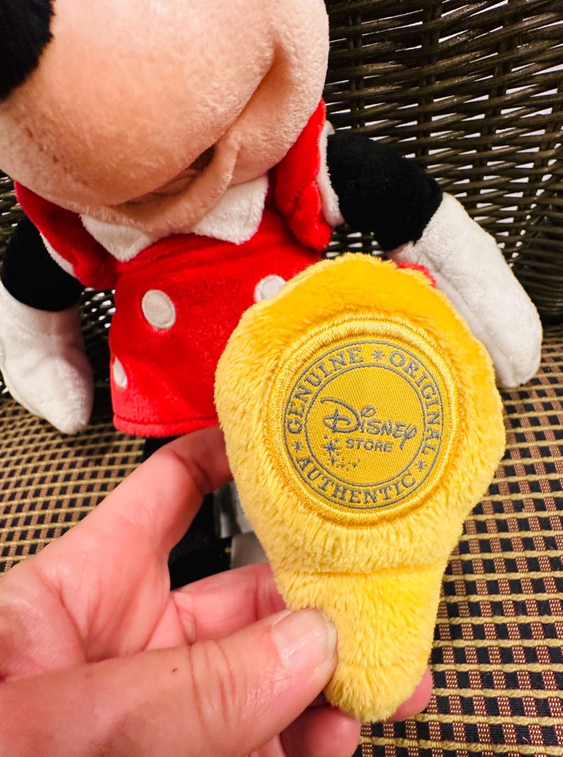 Puede incluir: Mu&ntilde;eca de peluche de Minnie Mouse con un vestido rojo con lunares blancos y un cuello blanco. Una etiqueta amarilla con el texto "GENUINE ORIGINAL Disney STORE AUTHENTIC" se sostiene frente a la mu&ntilde;eca.