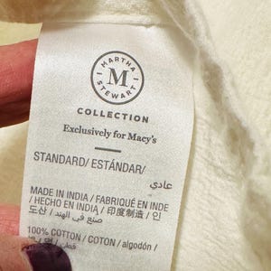 May include: Close-up of a white fabric tag with the text "MARTHA STEWART COLLECTION" and "Exclusively for Macy's". The tag also includes the text "STANDARD/EST&Aacute;NDAR" and "MADE IN INDIA". The fabric is a light cream color.