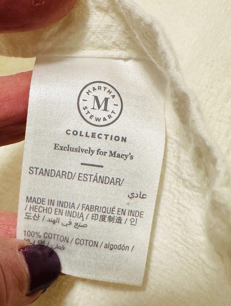 May include: Close-up of a white fabric tag with the text "MARTHA STEWART COLLECTION" and "Exclusively for Macy's". The tag also includes the text "STANDARD/EST&Aacute;NDAR" and "MADE IN INDIA". The fabric is a light cream color.