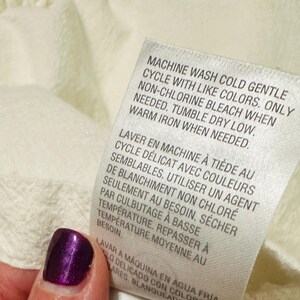 May include: Close-up of a white fabric tag with care instructions in English and French. The tag reads "MACHINE WASH COLD GENTLE CYCLE" and other washing instructions. A hand with a purple nail is holding the tag.