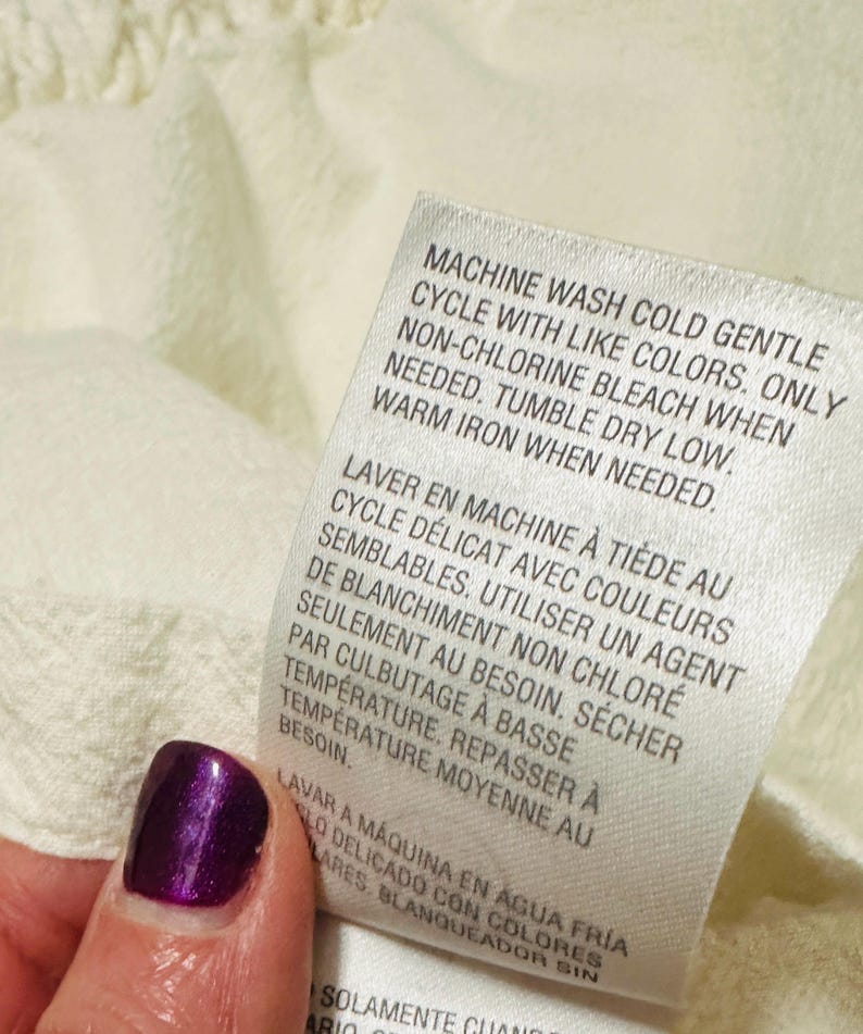 May include: Close-up of a white fabric tag with care instructions in English and French. The tag reads "MACHINE WASH COLD GENTLE CYCLE" and other washing instructions. A hand with a purple nail is holding the tag.