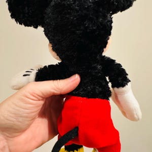 Puede incluir: Un peluche de Mickey Mouse, visto desde atr&aacute;s. El juguete presenta pelo negro, orejas grandes y redondas, pantalones cortos rojos, zapatos amarillos y guantes blancos. Se ve una cola negra. El juguete est&aacute; sostenido por una persona.