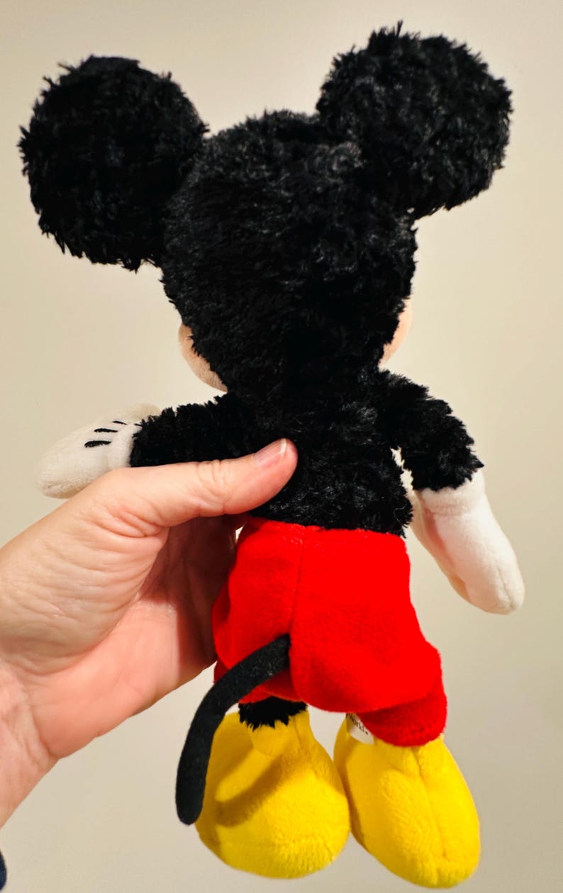 Puede incluir: Un peluche de Mickey Mouse, visto desde atr&aacute;s. El juguete presenta pelo negro, orejas grandes y redondas, pantalones cortos rojos, zapatos amarillos y guantes blancos. Se ve una cola negra. El juguete est&aacute; sostenido por una persona.