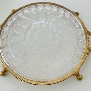May include: A clear glass serving tray with a decorative gold-colored metal rim and handles. The tray has a circular design with a central pattern and a series of petal-shaped indentations around the edge. The tray is supported by small, ornate feet.