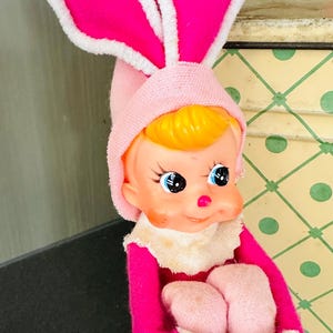 May include: A vintage Easter doll wearing a pink bunny costume with large ears and a matching bonnet. The doll has a painted face with blue eyes and yellow hair. The doll is sitting, with pink feet and hands.
