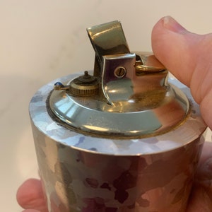 May include: A silver-toned, vintage lighter with a textured, hammered finish. The lighter has a hinged top and a small, circular dial. The lighter is held in a hand.