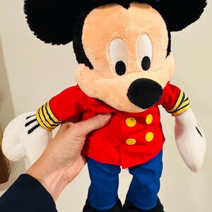 May include: A plush Mickey Mouse doll wearing a red jacket with yellow buttons, blue pants, and black shoes. The doll has a white sailor hat with a blue brim and the letters "DCL". The doll's ears are black, and its gloves are white.