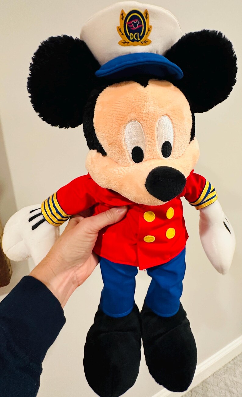 May include: A plush Mickey Mouse doll wearing a red jacket with yellow buttons, blue pants, and black shoes. The doll has a white sailor hat with a blue brim and the letters "DCL". The doll's ears are black, and its gloves are white.